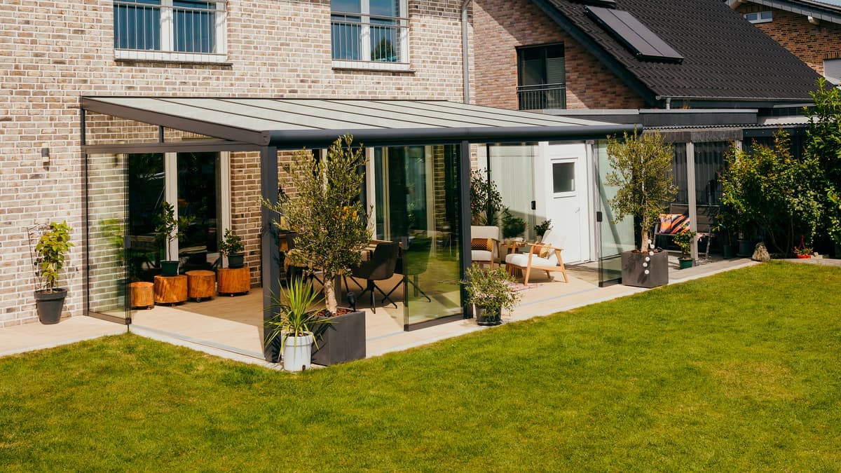 Terrasoverkapping aan een woning bij zonsondergang, met een stel dat ontspannen op een comfortabele bank zit.Véranda avec parois coulissantes en verre et éclairage d'ambiance
